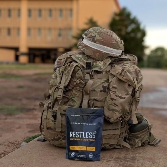 Military rucksack and helmet placed on the ground with RESTLESS® Performance Energy Blend in front—tactical fuel for soldiers and high-demand professionals